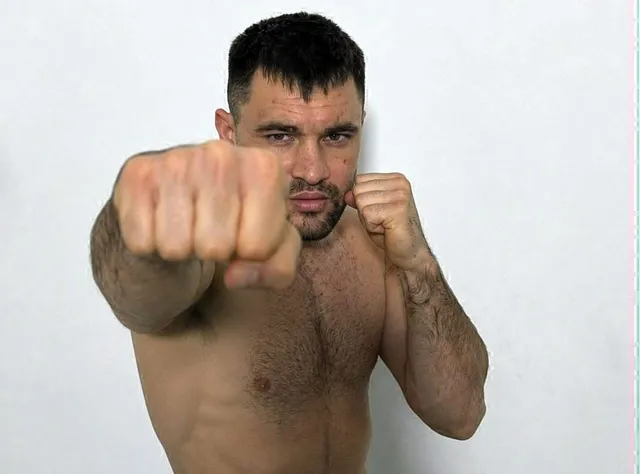 Un bărbat fără tricou stă în poziție de luptă cu pumnii strânși în fața unui fundal alb.