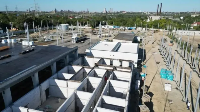 O vedere de sus prezintă un șantier cu structuri de beton, stâlpi metalici, un camion alb și utilaje pe un teren deschis sub cerul senin.