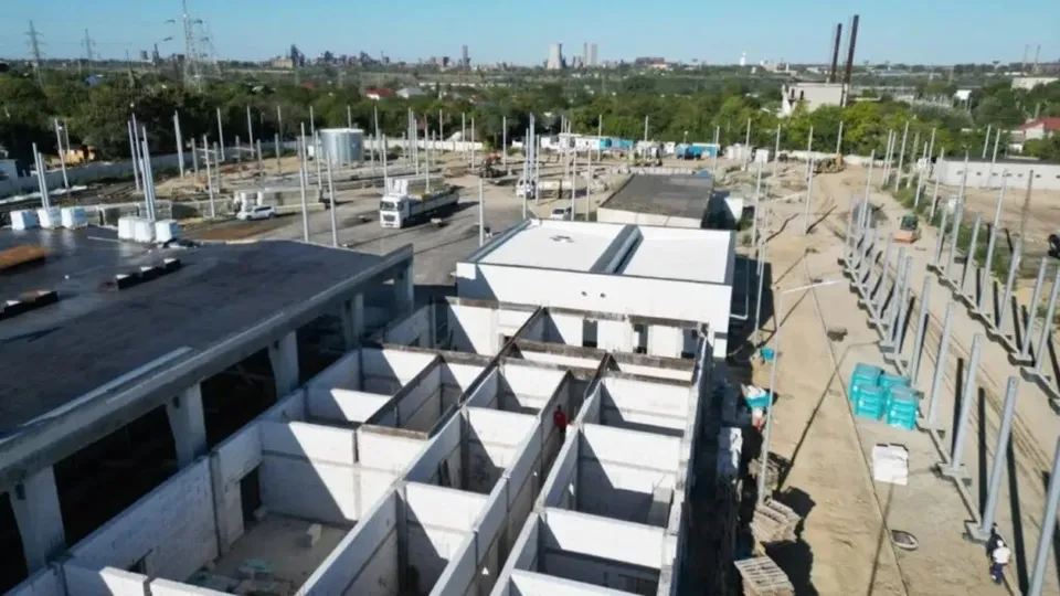 O vedere de sus prezintă un șantier cu structuri de beton, stâlpi metalici, un camion alb și utilaje pe un teren deschis sub cerul senin.