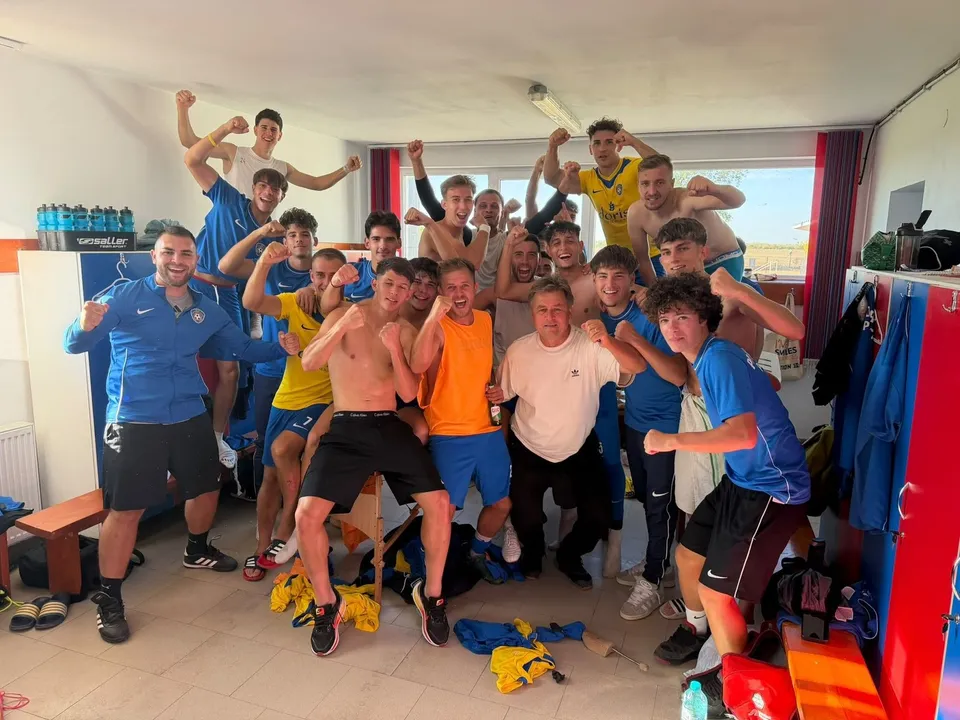 Un grup de tineri bărbați stau într-o cameră de vestiar, unii purtând tricouri colorate și pantaloni scurți, în timp ce alții sunt fără tricou.