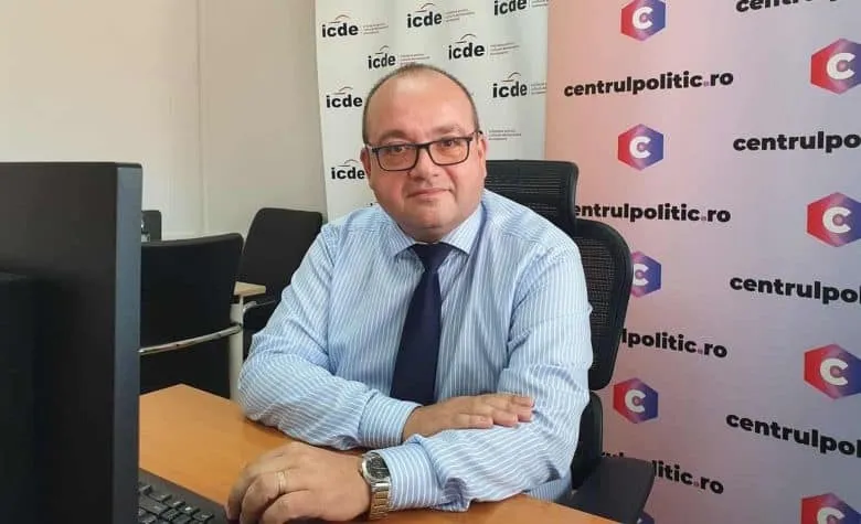 Un bărbat cu ochelari și cămașă albastră cu dungi stă la un birou lângă un monitor și un fundal cu logouri.