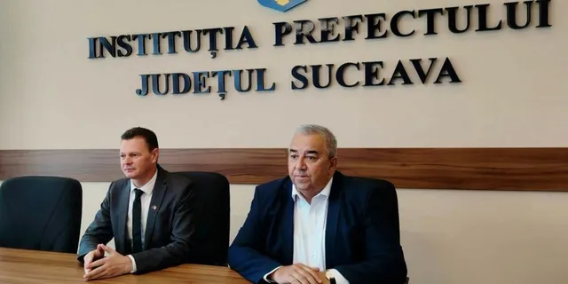 Două bărbați stau la o masă de lemn în fața unui perete pe care este scris un text în litere negre.