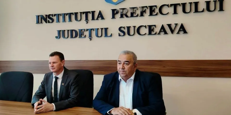 Două bărbați stau la o masă de lemn în fața unui perete pe care este scris un text în litere negre.