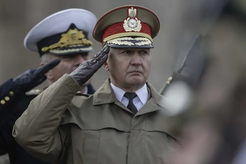 Un bărbat în uniformă militară cu șapcă decorată execută salutul militar în timp ce un alt bărbat cu șapcă albă stă în spatele lui.