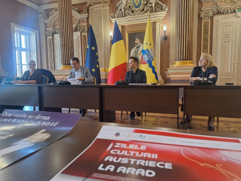 Patru persoane stau la o masă lungă într-o încăpere cu coloane, în fața unor steaguri și sub un portret încadrat.