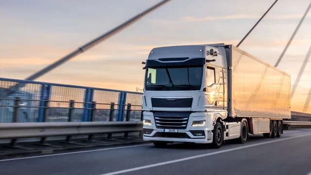 Un camion alb cu remorcă mare circulă pe o autostradă lângă un parapet metalic sub un cer la apus.
