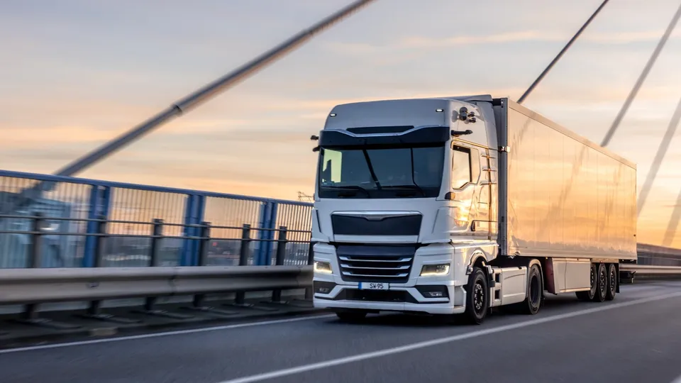 Un camion alb cu remorcă mare circulă pe o autostradă lângă un parapet metalic sub un cer la apus.