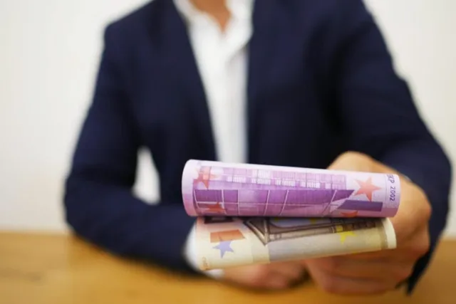 O persoană purtând sacou închis și cămașă albă ține două bancnote euro rulate pe o suprafață de lemn.