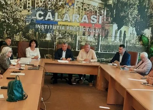 Șase persoane stau la o masă de conferință de lemn sub un poster mare cu textul CALARASI INSTITUTIA PREFECTULUI.