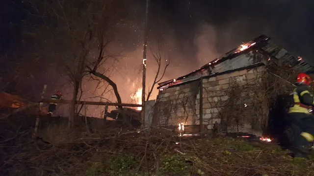 Flăcări și fum se ridică deasupra unor structuri de lemn și ziduri de cărămidă într-un peisaj nocturn, lângă un pompier cu echipament de protecție.