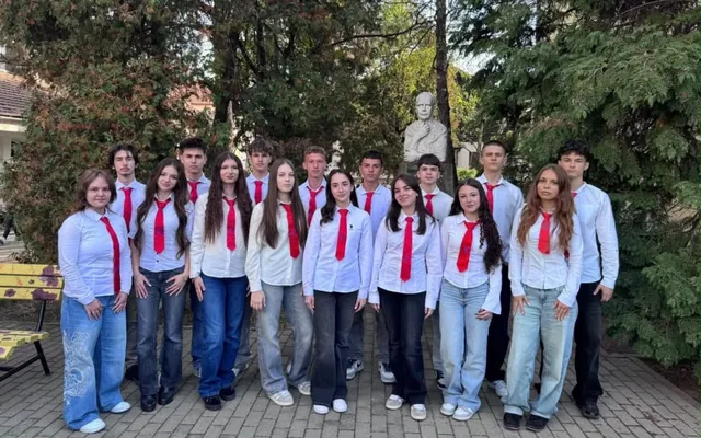 Un grup de elevi îmbrăcați în cămăși albe cu cravate roșie stau poziționați pe o curte cu copaci și un bust în fundal.