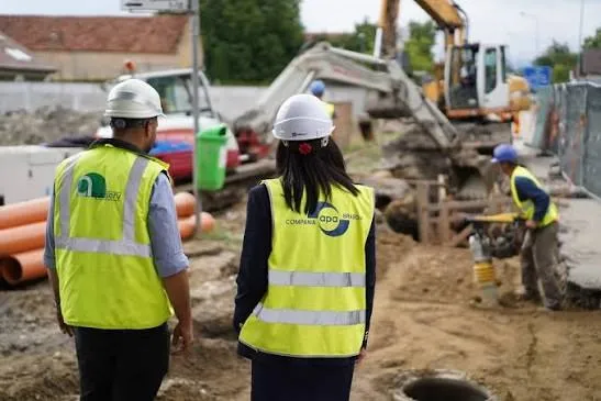 Două persoane purtând veste reflectorizante și căști de protecție privesc un șantier cu o excavatoare și țevi de culoare portocalie.