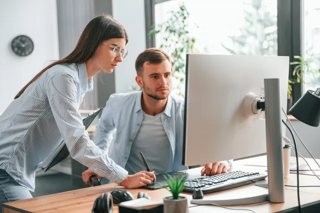 O femeie cu ochelari și o cămașă albastră stă lângă un bărbat care privește un monitor de calculator pe un birou de lemn.