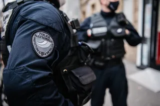 Un agent de poliție poartă o uniformă albastră cu un emblema pe braț, în timp ce un alt agent stă în fundal.