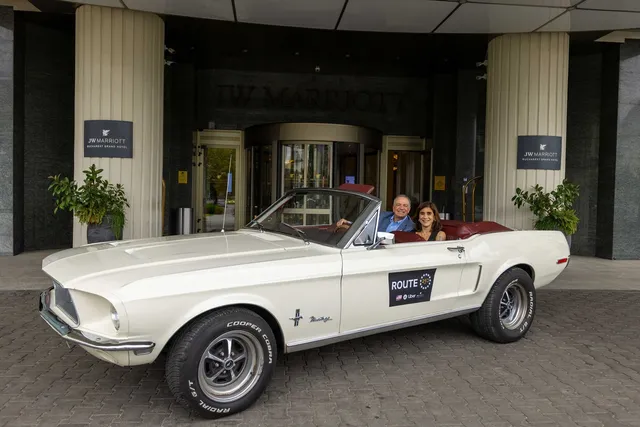 Un autoturism vintage alb cu decapotă este parcat în fața unei intrări de hotel, având două persoane așezate în interiorul acestuia.