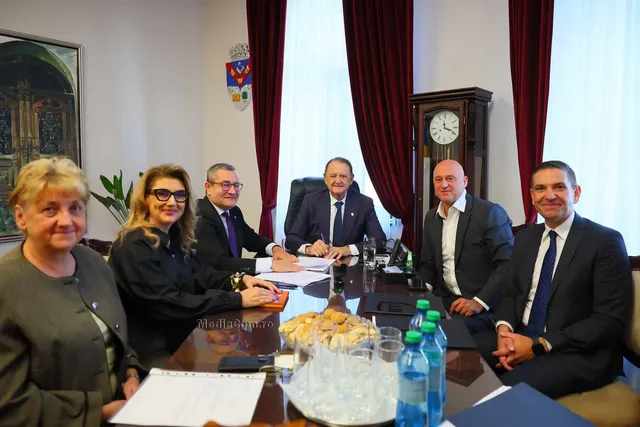 Șase persoane stau la o masă de conferințe într-o încăpere cu perdele roșii, având pe masă documente, apă și un coș cu produse de panificație.