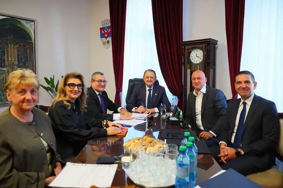Șase persoane stau la o masă de conferințe într-o încăpere cu perdele roșii, având pe masă documente, apă și un coș cu produse de panificație.