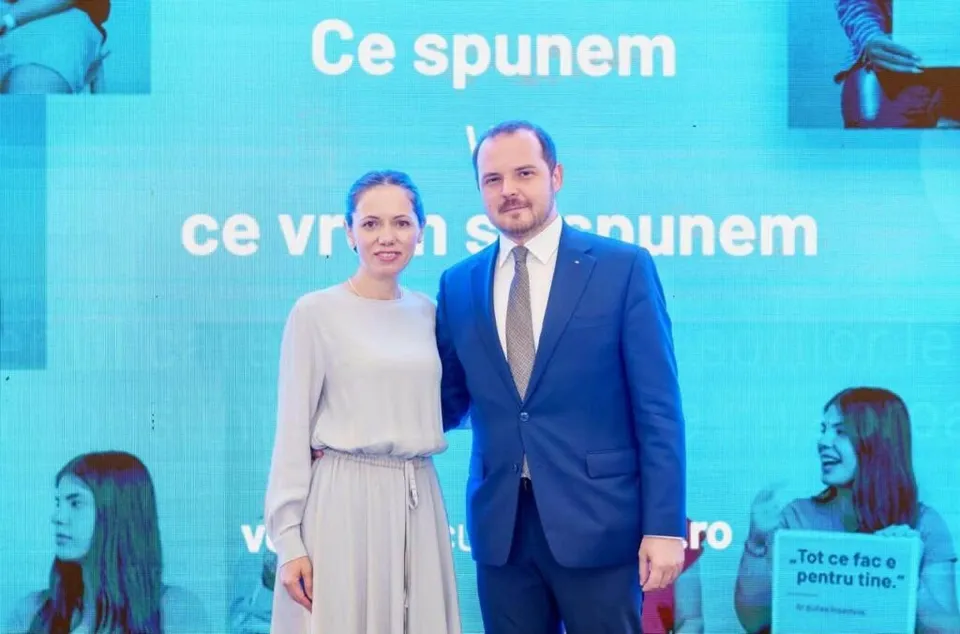 O femeie într-o rochie deschisă la culoare și un bărbat într-un costum albastru stau în picioare în fața unui ecran albastru cu text alb.