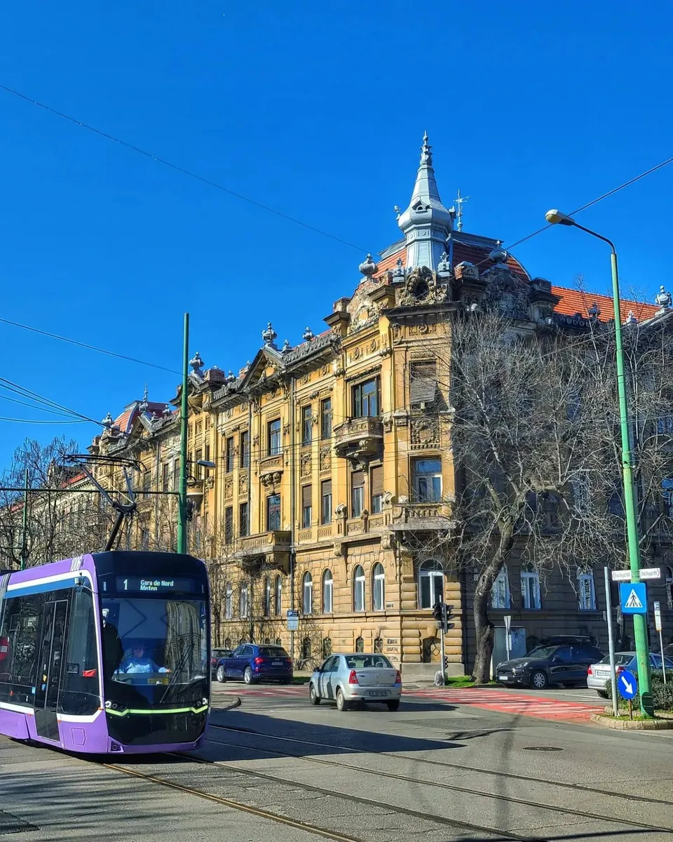Un tramvai mov trece pe o stradă lângă un clădire veche de culoare galbenă sub un cer senin.