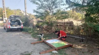 O persoană în haine roșii stă ghemuită lângă un panou informativ încadrat în lemn, pe un drum de țară lângă un autoturism argintiu.