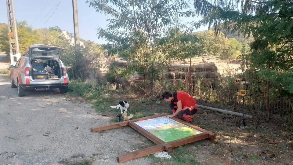 O persoană în haine roșii stă ghemuită lângă un panou informativ încadrat în lemn, pe un drum de țară lângă un autoturism argintiu.