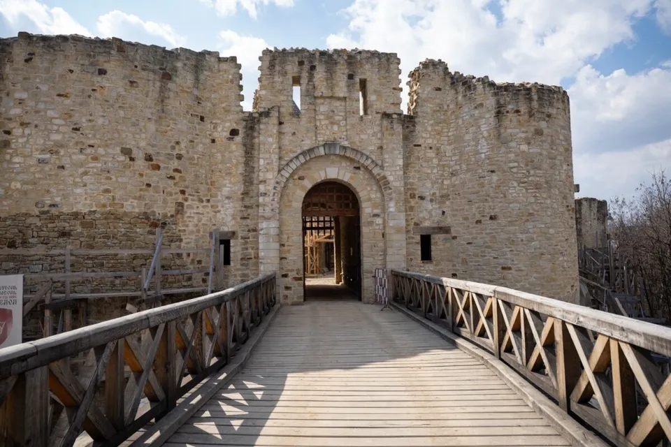 O punte de lemn cu balustrade din lemn conduce către o poartă de piatră dintr-un zid de fortificație sub un cer cu nori.