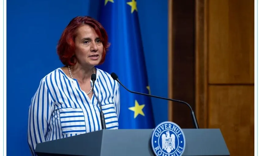 O femeie cu părul roșu, purtând o cămașă albastră cu dungi albe, stă în spatele unui podium cu un emblema oficială.