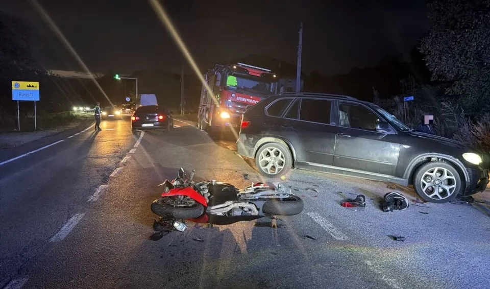 O motocicletă roșie și neagră este proiectată pe asfalt în timp ce un SUV gri închis este oprit pe partea laterală a drumului, într-o scenă de accident nocturnă.