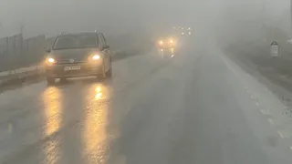 O mașină cu farurile aprinse parcurge o șosea wet și încețoșată, cu vizibilitatea redusă din cauza ceții dense.