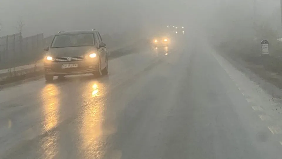 O mașină cu farurile aprinse parcurge o șosea wet și încețoșată, cu vizibilitatea redusă din cauza ceții dense.