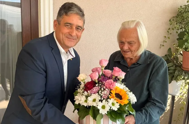 Un bărbat într-un costum albastru și o femeie în vârstă stau lângă un buchet de flori colorate.
