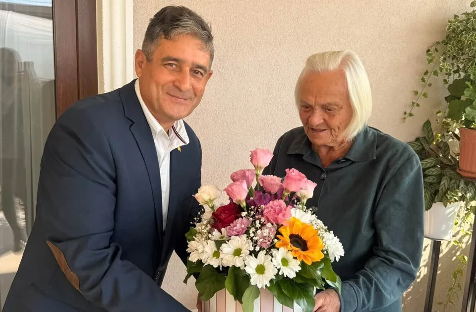 Un bărbat într-un costum albastru și o femeie în vârstă stau lângă un buchet de flori colorate.
