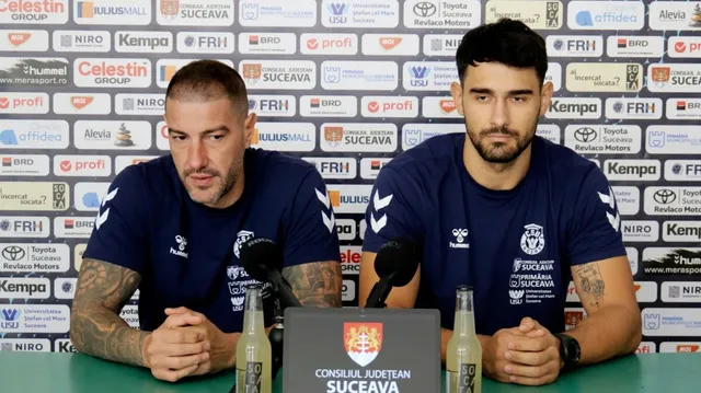Doi bărbați cu tricouri albastre de sport stau la o masă cu microfoane și sticle de apă în fața unui panou cu diverse logo-uri.