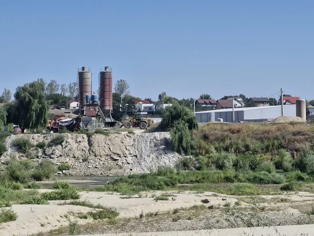 Două silozuri înalte și un utilaj de construcții sunt situate lângă un perete de pământ și pietriș, sub un cer senin.