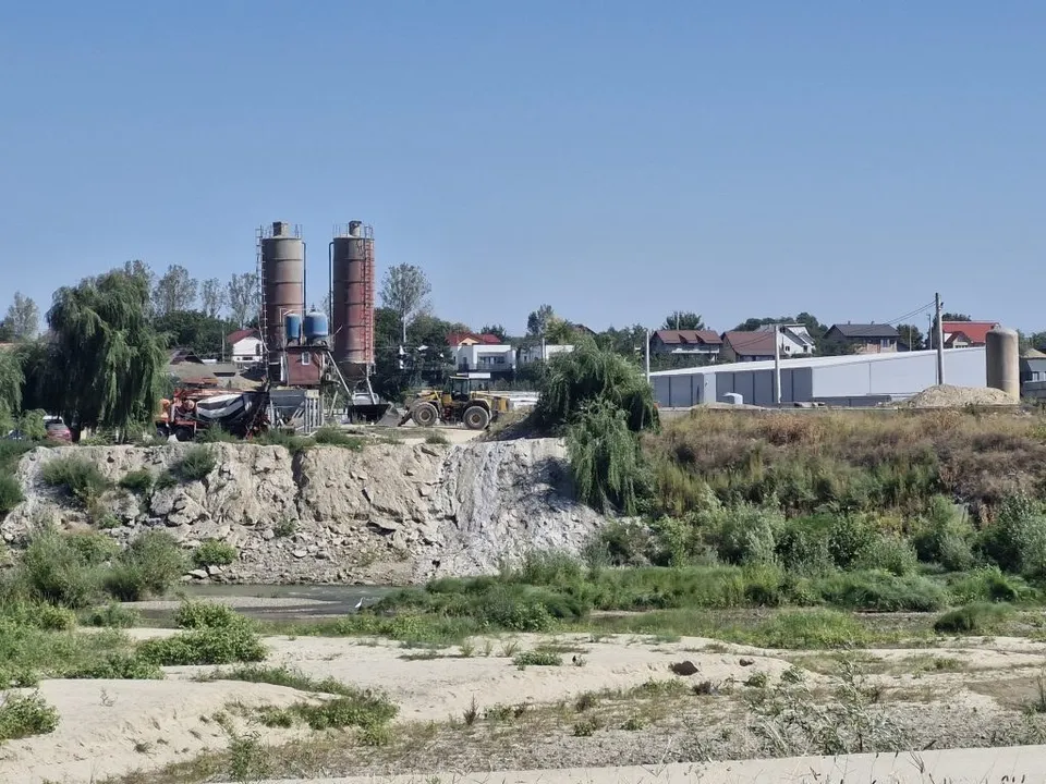 Două silozuri înalte și un utilaj de construcții sunt situate lângă un perete de pământ și pietriș, sub un cer senin.