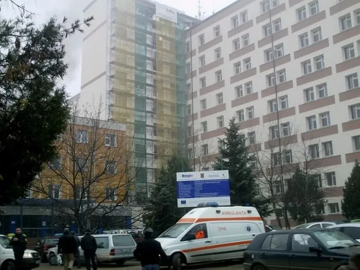Un vehicul de tip ambulanță alb cu detalii roșii este parcat în fața unor clădiri înalte de beton lângă câțiva oameni și mașini.