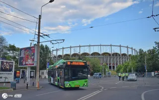 Un autobuz verde și negru circulă pe un drum asfaltat în fața unei structuri circulare mari sub un cer parțial înzăgoțat.