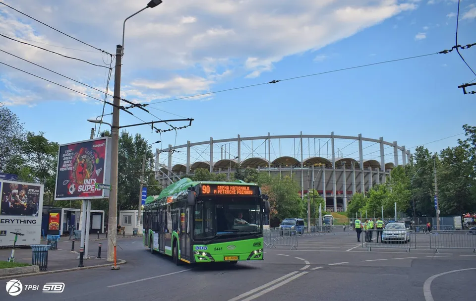 Un autobuz verde și negru circulă pe un drum asfaltat în fața unei structuri circulare mari sub un cer parțial înzăgoțat.