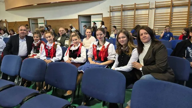 Un grup de persoane, inclusiv copii în costume tradiționale și adulți, stau așezați pe scaune albastre într-o sală cu panouri de lemn pe perete.