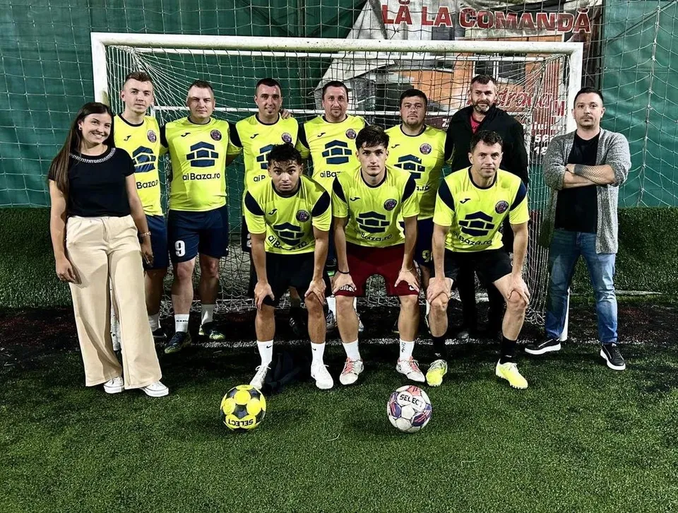 O echipă de fotbal masculină în echipamente galben și negru pozează lângă un gol, alături de o femeie și un bărbat pe un teren de fotbal.
