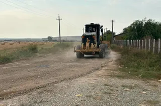 Un tractor cu o lamă frontală parcurge un drum de țară prăfuit lângă un câmp și un gard de beton.