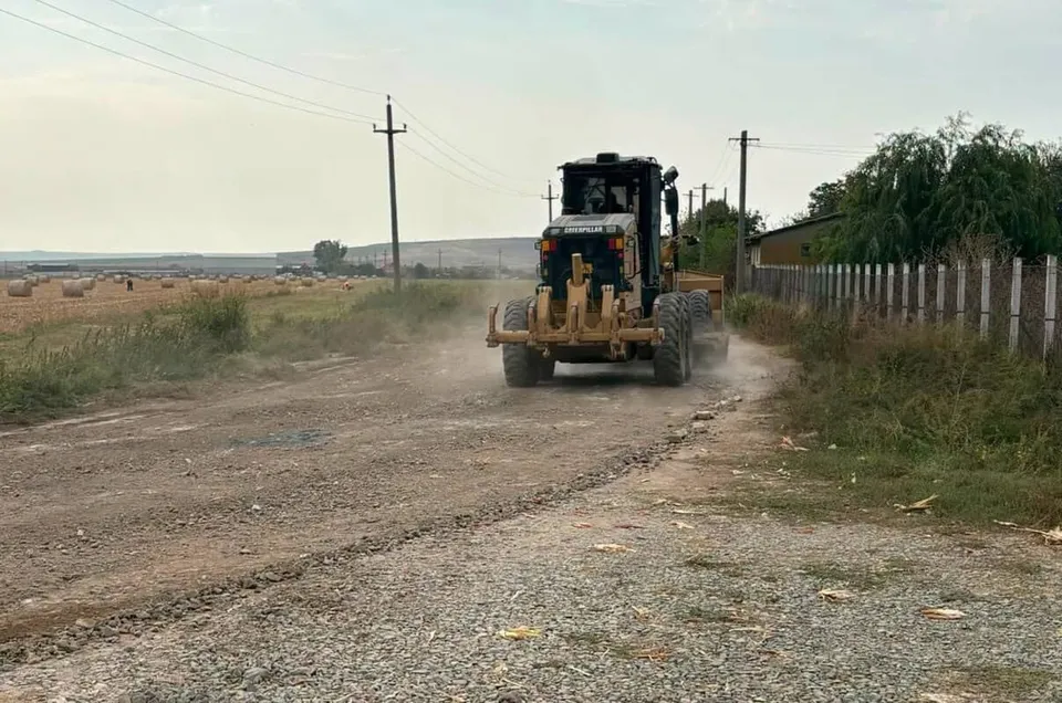 Un tractor cu o lamă frontală parcurge un drum de țară prăfuit lângă un câmp și un gard de beton.