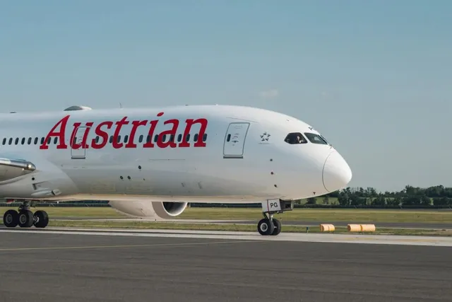Un avion de pasageri alb cu inscripția roșie Austrian se află pe o pistă de aterizare sub un cer senin.