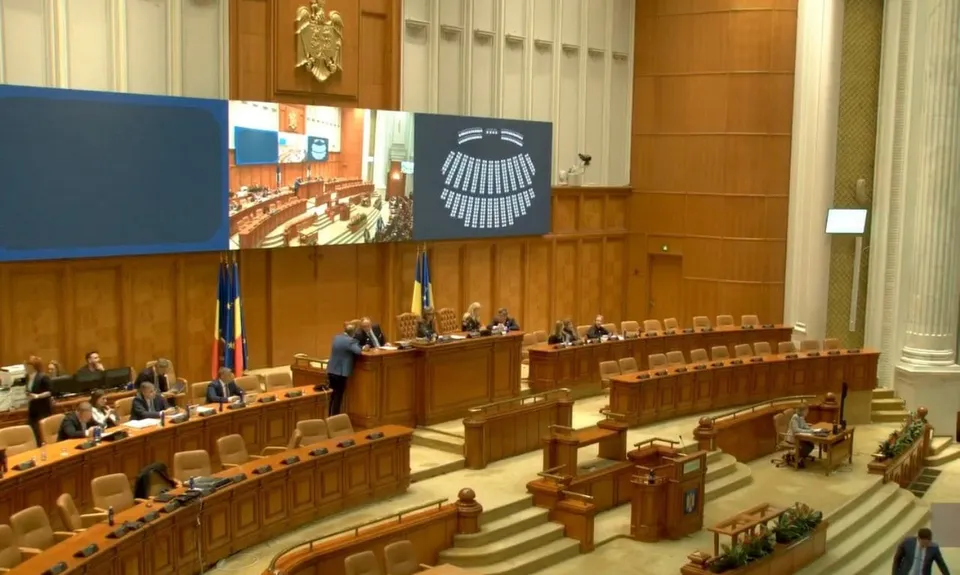 O sală de ședințe cu mese de lemn dispuse în rânduri, oameni așezați la birouri și ecrane mari montate pe un perete de lemn.