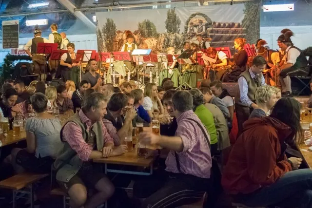 Oamenii stau la mese de lemn într-o încăpere mare, în timp ce un grup de muzicieni cântă pe o scenă în fundal.