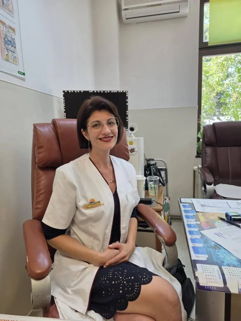O femeie cu ochelari și halat alb stă așezată într-un fotoliu maro într-un birou medical.