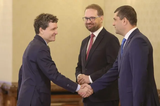 Trei bărbați în costume purtând cămăși albe stau în picioare și doi dintre aceștia se strâng în mână.