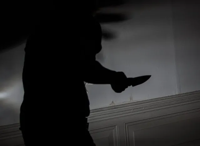 Silueta unei persoane întinse care ține un obiect tăietor în mână în fața unui perete deschis la culoare.