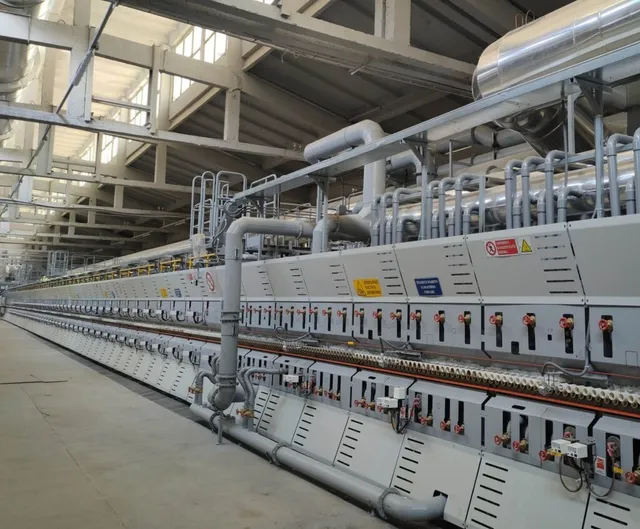 O linie lungă de echipamente industriale gri cu diverse țevi, cabluri și panouri de control este instalată într-un spațiu de depozitare mare.