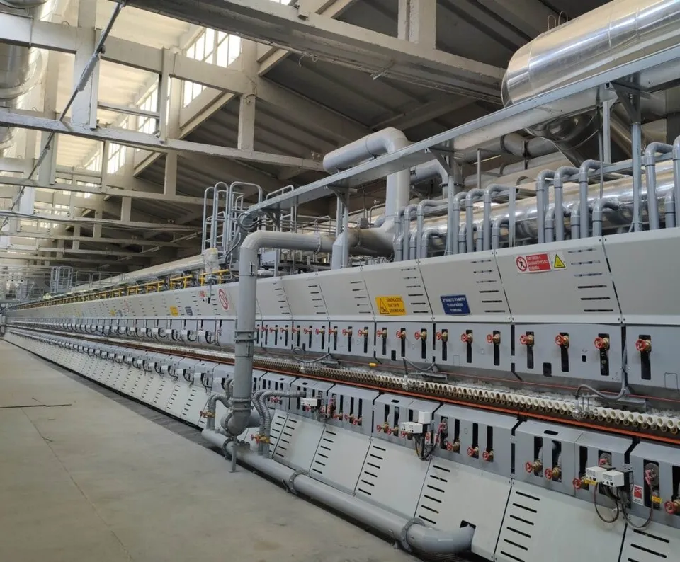 O linie lungă de echipamente industriale gri cu diverse țevi, cabluri și panouri de control este instalată într-un spațiu de depozitare mare.